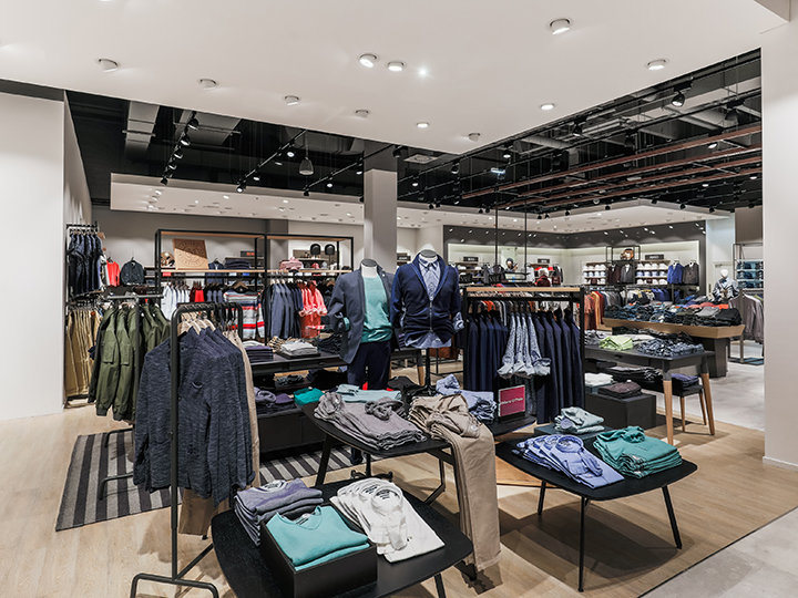 Ein modernes Interieur eines Bekleidungsgeschäfts mit ordentlich angeordneten Regalen und Tischen mit verschiedenen Kleidungsstücken wie Hemden, Jacken und Hosen. Der Raum ist mit Scheinwerfern gut beleuchtet und hat ein sauberes, organisiertes Layout mit neutralen Tönen.