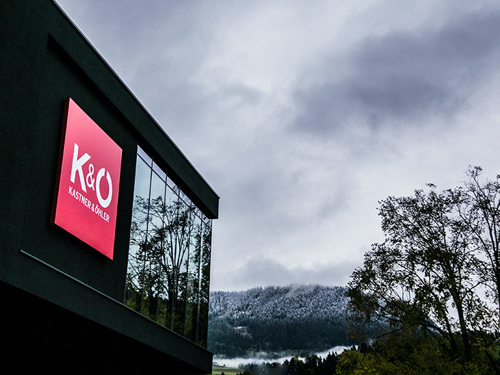 Vor einem wolkigen Himmel ist ein modernes Gebäude mit einem großen roten Schild mit der Aufschrift „K&O Kastner & Öhler“ abgebildet. Schneebedeckte Hügel und belaubte Bäume umgeben das Gebäude und schaffen einen Kontrast zwischen städtischen und natürlichen Elementen.