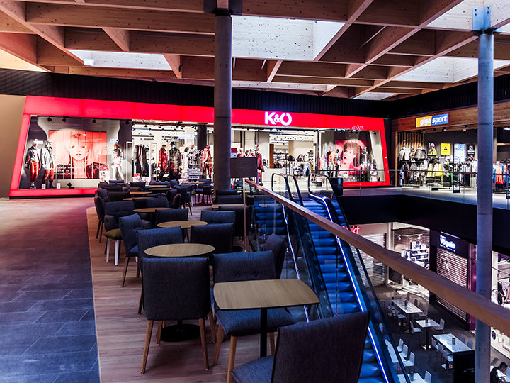 Das Interieur eines modernen Einkaufszentrums mit einem rot beleuchteten Bekleidungsgeschäft namens K&O. Der Raum umfasst Sitzbereiche mit Stühlen und Tischen, eine Rolltreppe und verschiedene andere Schaufenster im Hintergrund.