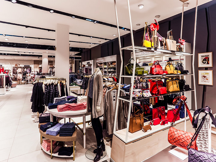 Ein Bekleidungs- und Accessoiregeschäft mit Regalen voller bunter Handtaschen, Schals und ordentlich gefalteter Kleidung. Schaufensterpuppen tragen modische Outfits. Das Geschäft ist hell beleuchtet und modern eingerichtet.