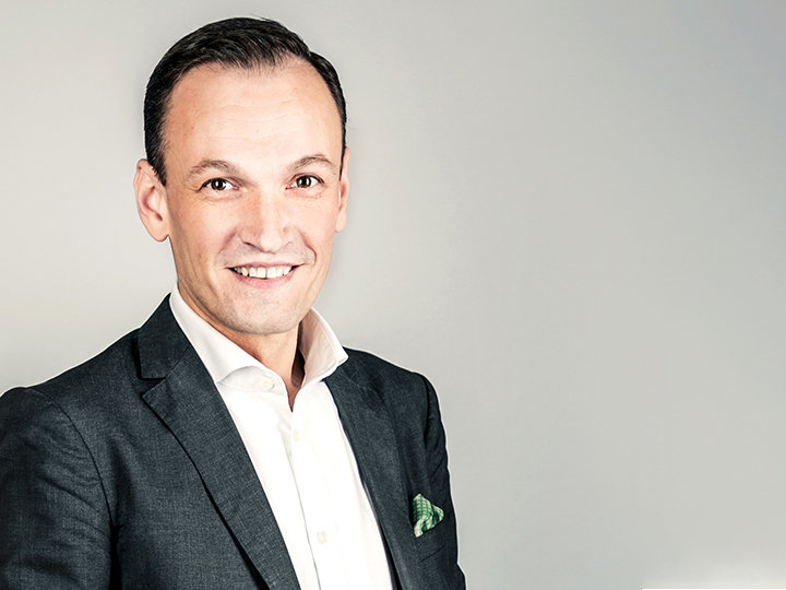 Ein Mann in dunklem Anzug und weißem Hemd lächelt vor einem hellgrauen Hintergrund. Er hat kurze Haare und ein Einstecktuch in seinem Jackett.