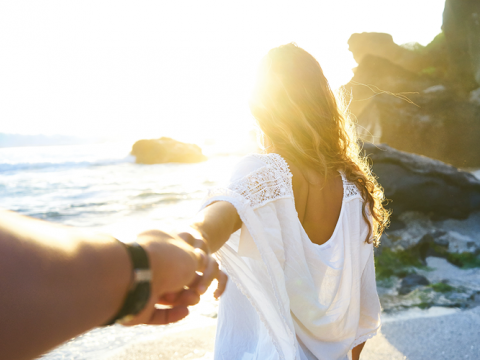Eine Person in einem weißen Kleid geht an einem sonnigen Strand entlang und hält dabei die Hand einer anderen Person. Im Hintergrund sind das Meer und große Felsen zu sehen, und das Sonnenlicht erzeugt eine warme, leuchtende Atmosphäre.