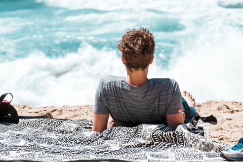 Eine Person mit kurzen Haaren und einem grauen T-Shirt liegt auf einem gemusterten Handtuch am Strand. Sie blickt auf das Meer, im Hintergrund brechen die Wellen.
