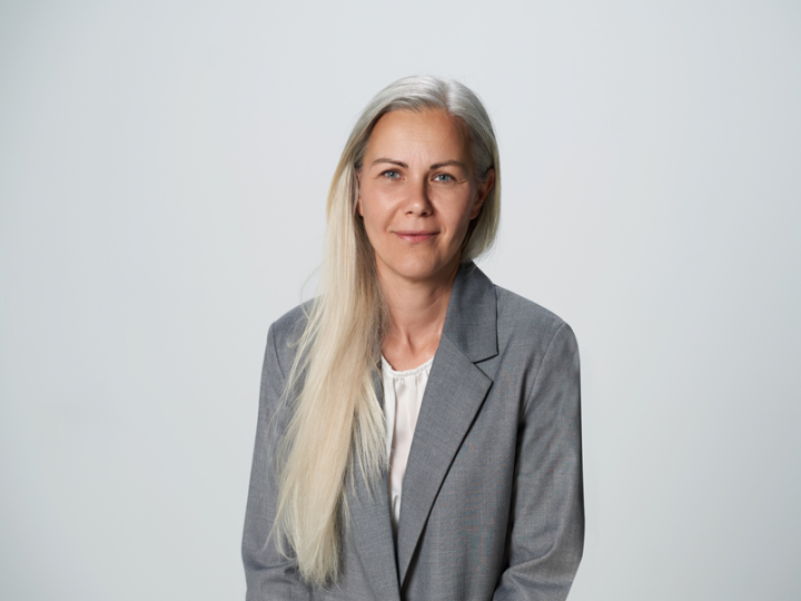 Eine Person mit langen blonden Haaren, einem hellgrauen Blazer und einer weißen Bluse steht vor einem schlichten, hellgrauen Hintergrund. Sie hat einen neutralen Gesichtsausdruck und blickt in die Kamera.