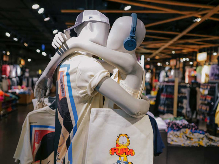 Zwei Schaufensterpuppen stehen in einem Bekleidungsgeschäft umarmt. Eine trägt eine Mütze und ein T-Shirt mit Aufdruck, die andere trägt Kopfhörer und eine Einkaufstasche mit einem Katzen-Cartoon-Design. Im Hintergrund sind verschiedene Kleidungsstücke und hölzerne Deckenbalken zu sehen.