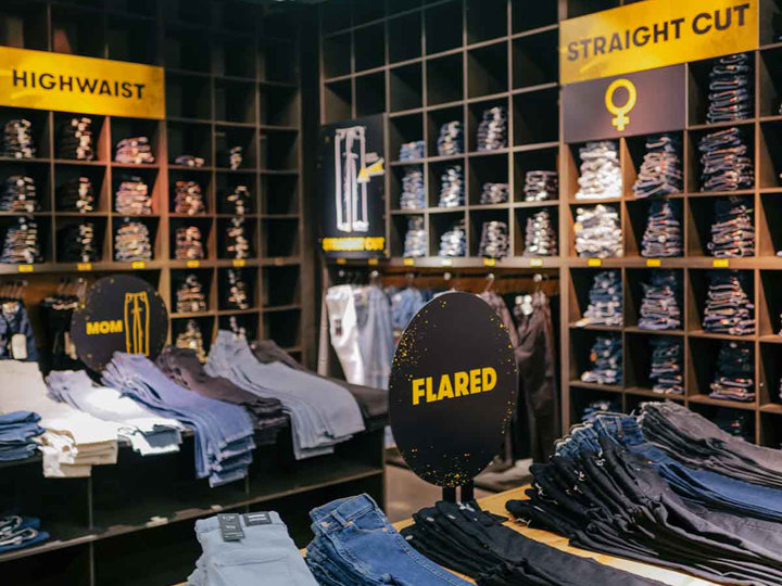 In einem Bekleidungsgeschäft werden verschiedene Jeansstile präsentiert, darunter High Waist, Straight Cut, Mom und Flared. Die Jeans sind ordentlich auf Holzregalen und Tischen gestapelt. Helle Schilder weisen auf die verschiedenen verfügbaren Stile hin.