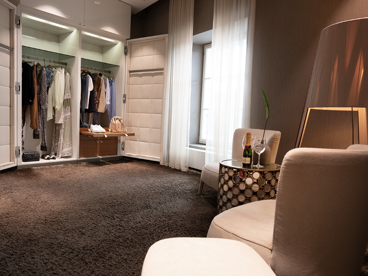 Ein modernes, elegantes Zimmer mit einem offenen Kleiderschrank, in dem Kleidung und eine Handtasche ausgestellt sind. Zwei bequeme Stühle stehen vor einem runden Tisch mit einer dekorativen Lampe. Eine Champagnerflasche und Gläser stehen auf dem Tisch und Licht strömt durch die transparenten Vorhänge.