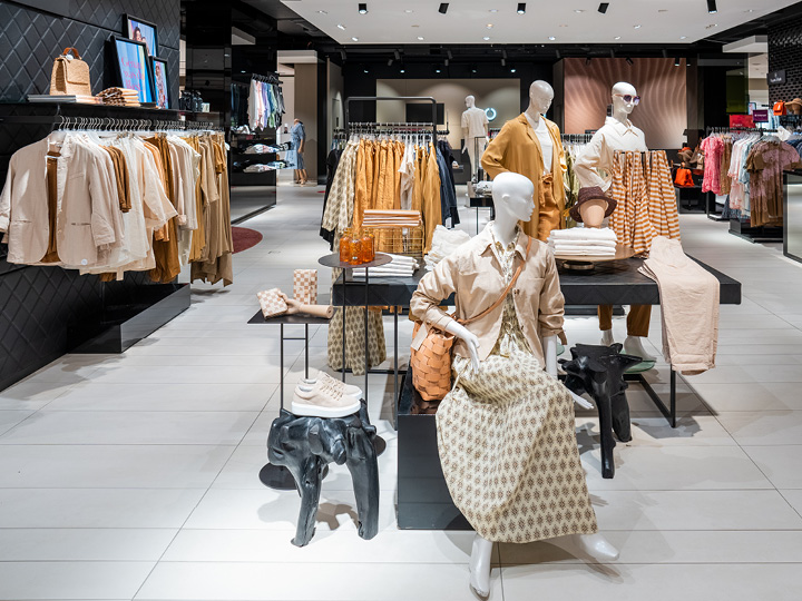 Inneneinrichtung eines Bekleidungsgeschäfts mit Kleidung in neutralen Farbtönen auf Ständern und Schaufensterpuppen. Ausgestellt werden Hemden, Jacken, Kleider und Accessoires wie Taschen und Hüte. Hell beleuchtet, mit modernem, übersichtlichem Layout.