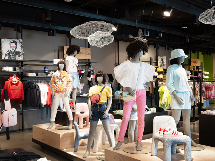 In einem trendigen Bekleidungsgeschäft sind Schaufensterpuppen in farbenfrohen, legeren Outfits zu sehen. Das Geschäft bietet eine Vielzahl von Kleidungsstücken und Accessoires vor einem stilvollen, modernen Hintergrund mit künstlerischen Dekorelementen, die von der Decke hängen.