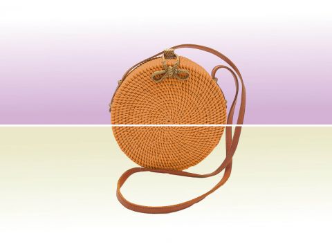 Eine runde, gewebte Handtasche mit einem braunen Lederriemen ruht auf einem horizontal geteilten Hintergrund, in dem oben Hellviolett und unten Blassgrün zu sehen ist, wodurch ein harmonischer Kontrast entsteht.