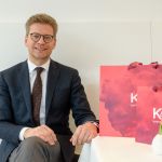Ein Mann in Anzug und Brille sitzt lächelnd mit einer roten Einkaufstüte an einem Tisch. Auf dem Tisch steht auch eine kleine weiße Keramikvase.