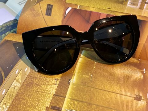 Eine übergroße schwarze Sonnenbrille mit dunklen Gläsern liegt auf einer glänzenden, goldfarbenen Oberfläche. Der Hintergrund zeigt Text und verschwommene architektonische Elemente.