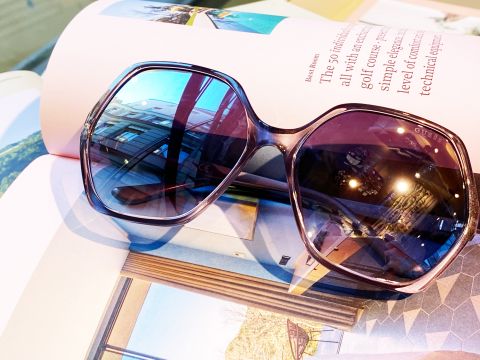 Große Sonnenbrillen mit violetten Gläsern liegen auf einer aufgeschlagenen Zeitschrift, die Bilder von Architektur und Innenräumen zeigt. Die Sonnenbrillen reflektieren einige der Deckenleuchten und das Dekor des Raums.