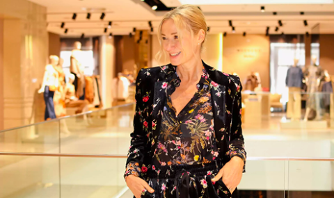 Eine Person mit blondem Haar steht lächelnd in einem hell erleuchteten Innenraum. Sie trägt ein Outfit mit Blumenmuster. Der Hintergrund ist leicht verschwommen und zeigt Kleidungsdisplays und andere Personen.