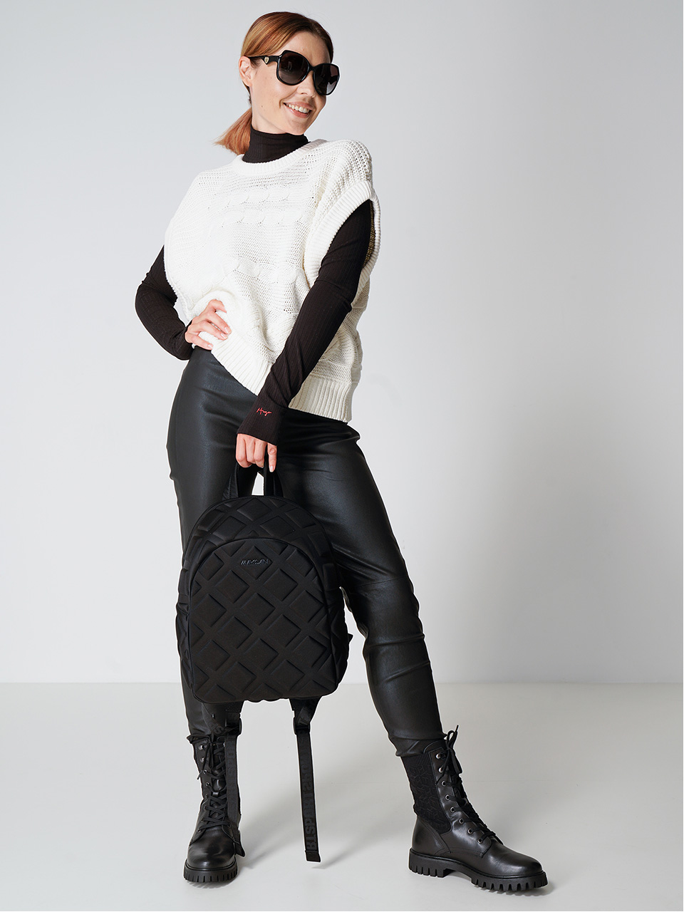 Eine stylische Frau mit schwarzer Sonnenbrille, einem weißen ärmellosen Pullover über einem schwarzen Rollkragenpullover und schwarzen Lederhosen posiert selbstbewusst. Sie hält einen schwarzen Rucksack und trägt schwarze Schnürstiefel. Sie steht vor einem schlichten Hintergrund.