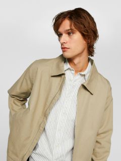 Eine Person mit mittellangem braunem Haar trägt eine hellbeige Jacke über einem weiß-grau gestreiften Hemd. Sie blickt zur Seite, hat eine Hand in der Tasche und steht vor einem schlichten weißen Hintergrund.