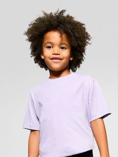 Ein kleines Kind mit lockigem Haar lächelt und trägt ein hellviolettes T-Shirt. Der Hintergrund ist schlicht und hell.