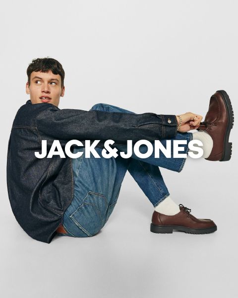 Mann sitzt auf dem Boden und trägt eine blaue Jeansjacke, Jeans und braune Schuhe. Er blickt zur Seite, hat die Knie angewinkelt und verschränkt die Arme vor den Knien. Der Text „JACK & JONES“ ist über das Bild gelegt.