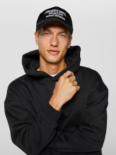 Mann trägt einen schwarzen Kapuzenpullover und eine schwarze Kappe mit weißem Text. Er hat einen neutralen Gesichtsausdruck und berührt den Kapuzenpullover mit seiner linken Hand. Der Hintergrund ist schlicht weiß.