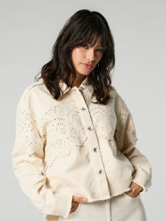 Eine Person mit langen dunklen Haaren trägt eine cremefarbene Jacke mit Knöpfen und Broderie-Anglaise-Details. Sie hat die Hände in den Taschen und steht vor einem neutralen Hintergrund.