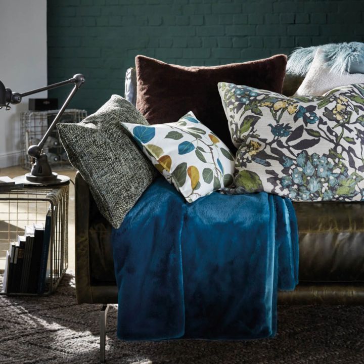 Eine gemütliche Wohnzimmerszene mit einem dunklen Ledersofa, das mit verschiedenen gemusterten Kissen und einer blaugrünen Decke geschmückt ist. Neben dem Sofa steht eine moderne schwarze Lampe, auf einem Beistelltisch stehen Bücher. Im Hintergrund befindet sich eine dunkelgrüne Ziegelwand.
