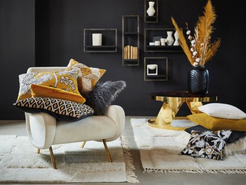 Ein modernes Wohnzimmer mit einem beigen Sessel, geschmückt mit verschiedenen gemusterten Kissen in Gelb-, Grau- und Weißtönen. Auf einem Couchtisch mit goldenen Beinen stehen eine schwarze Vase und Trockenpflanzen. An den dunklen Wänden hängen geometrische Regale mit Dekoartikeln.