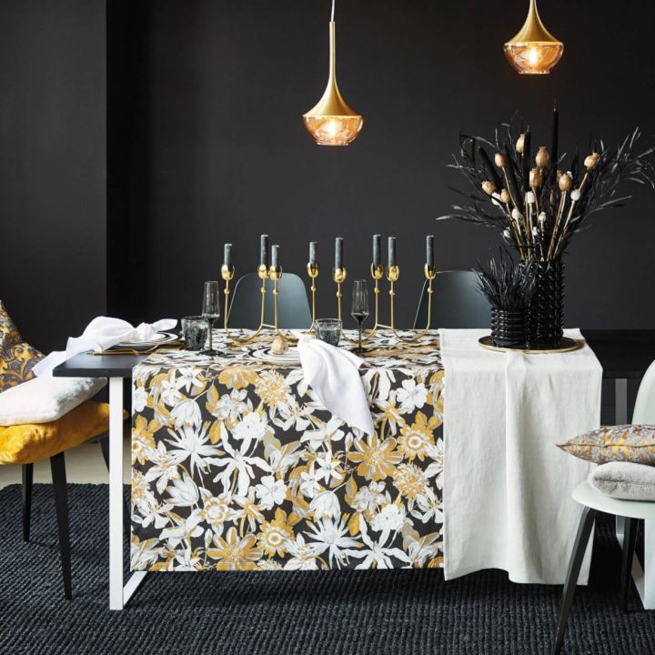 Ein modernes Esszimmer mit einem schwarzen Tisch und einer geblümten Tischdecke in Gelb und Weiß, geschmückt mit goldenen Kerzenständern und Glaswaren. Auf den Stühlen liegen dekorative Kissen, und der Hintergrund ist dunkel mit goldenen Hängelampen.