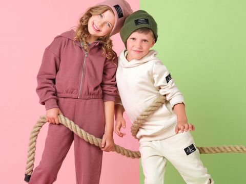 Zwei Kinder lächeln und halten ein Seil vor einem gespaltenen rosa und grünen Hintergrund. Das Mädchen trägt einen rosa Trainingsanzug und eine passende Mütze, während der Junge einen weißen Trainingsanzug und eine grüne Mütze trägt. Beide Kinder haben Markenaufnäher auf ihren Outfits.