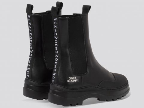 Schwarze Stiefeletten mit dicker Sohle und Laschen an der Rückseite. Auf den Laschen ist vertikal das Wort „WOMS“ aufgedruckt, und auf der Ferse ist „CHOOSE THE CHANGE“ aufgedruckt. Die Stiefel sind auf einem hellgrauen Hintergrund abgesetzt.