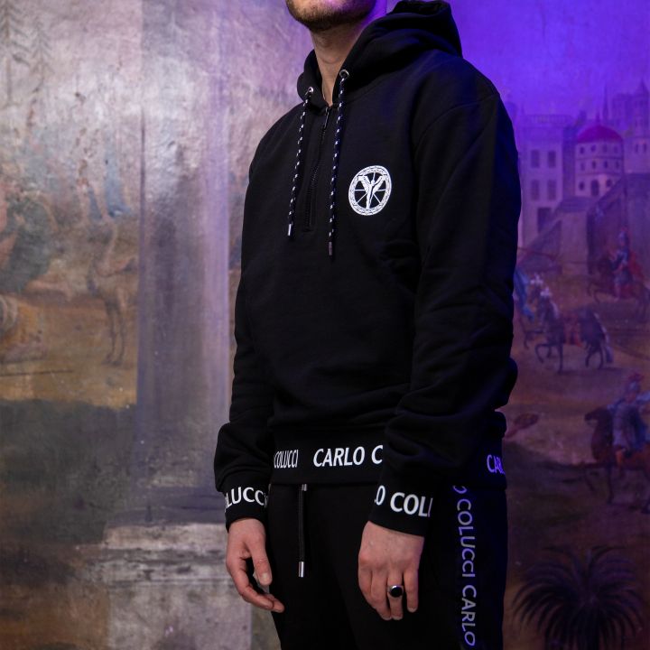 Ein Mann in einem schwarzen Kapuzenpullover und einer Hose mit dem Schriftzug „Carlo Colucci“ steht vor einem Wandgemälde mit klassischer Architektur, das teilweise mit violettem Licht beleuchtet ist. Er trägt eine schwarze Beanie und hat eine selbstbewusste Pose.