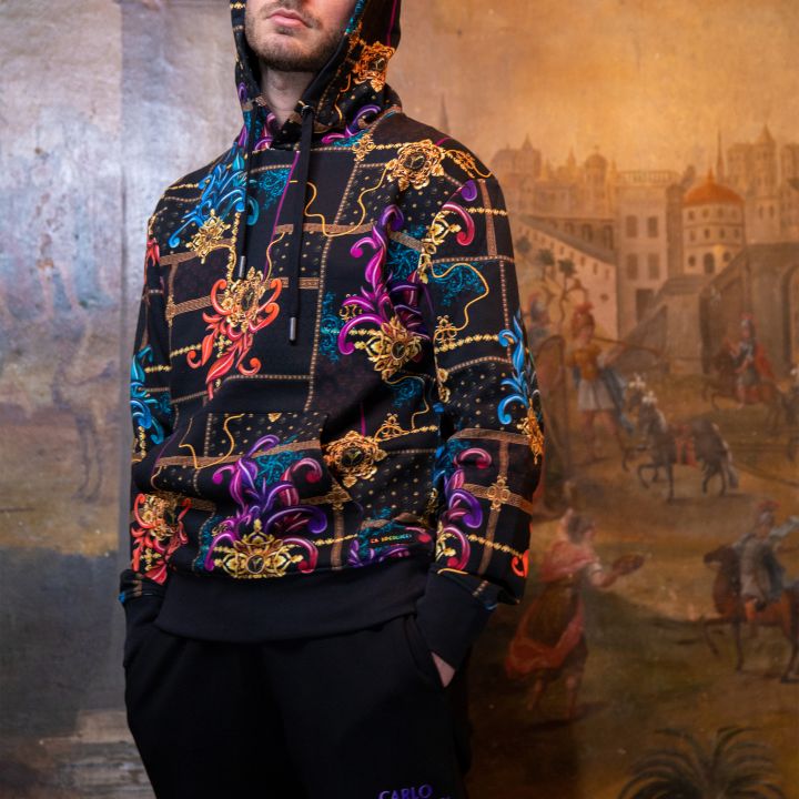 Eine Person mit einem bunt gemusterten Kapuzenpullover und schwarzen Hosen steht selbstbewusst mit den Händen in den Taschen. Im Hintergrund ist ein historisches Gemälde mit Gebäuden und Personen im klassischen Stil zu sehen.