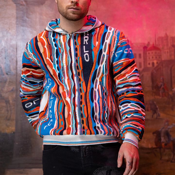 Ein Mann mit kurzen Haaren trägt einen bunten, gemusterten Pullover und schwarze Jeans. Er steht vor einem Wandbild in einer Mischung aus Rosa- und Orangetönen, das historische Architektur und eine Säule zeigt.