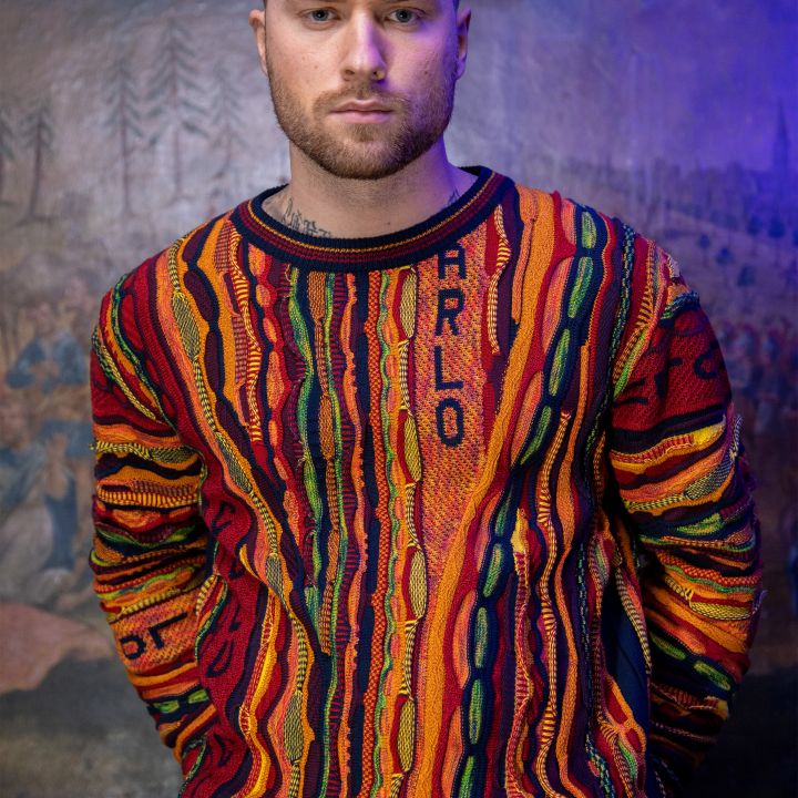 Vor einer gemalten Kulisse steht ein Mann mit kurzen Haaren und Bart. Er trägt einen bunten, gemusterten Pullover mit dem Wort „ARLO“ darauf. Der Hintergrund ist ein dunkles Wandgemälde im Vintage-Stil mit abstrakten Figuren.