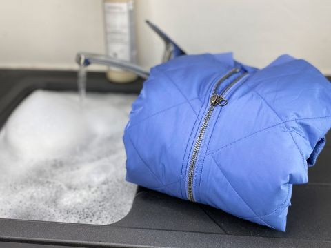 Eine blaue Jacke liegt auf dem Rand eines mit Seifenlauge gefüllten Waschbeckens, im Hintergrund läuft ein Wasserhahn.