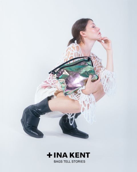 Auf einem weißen Hintergrund hockt eine Person, gekleidet in ein weißes Spitzenoutfit und schwarze Stiefel. Sie hält eine glänzende, bunte Handtasche in der Hand. Unten ist der Text „+ INA KENT BAGS TELL STORIES“ zu sehen.