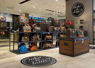 Ein Luxus-Ladendisplay, das verschiedene Ledertaschen und Koffer mit einem unverwechselbaren Monogrammmuster präsentiert. Der elegante Innenraum verfügt über Schaufensterpuppen und Regale mit Produkten. Im Vordergrund ist ein großer Koffer auffällig dargestellt.