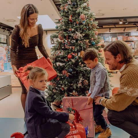 Eine Familie schmückt einen Weihnachtsbaum. Eine Frau hält ein Geschenk, während ein Mann kniet und zwei Kinder anlächelt. Das Mädchen sitzt auf einem roten Spielzeug und der Junge hält ein eingepacktes Geschenk. Der Baum ist mit Lichtern und Ornamenten geschmückt und steht in einem warm beleuchteten Raum.