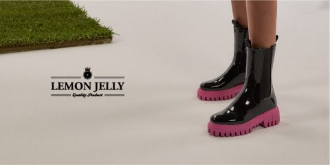 Person in glänzenden schwarzen Stiefeln mit rosa Sohlen auf weißem Boden, neben einem Fleck grünem Gras. Links steht der Text „Lemon Jelly Quality Product“.