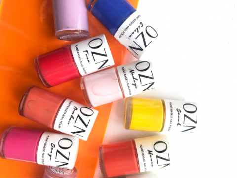 Eine Sammlung von neun Nagellackflaschen auf einer orangefarbenen Oberfläche. Die Farben umfassen Schattierungen von Rosa, Rot, Gelb, Blau, Lila und Weiß. Jede Flasche trägt die Aufschrift „OZN“.