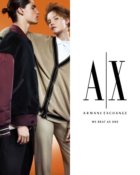 Zwei Models in stylischen Outfits posieren selbstbewusst vor einem orangefarbenen Hintergrund. Eine trägt eine dunkle Jacke, die andere eine hellbraune Strickjacke. Das Armani Exchange-Logo und der Slogan „We Beat As One“ sind rechts prominent zu sehen.