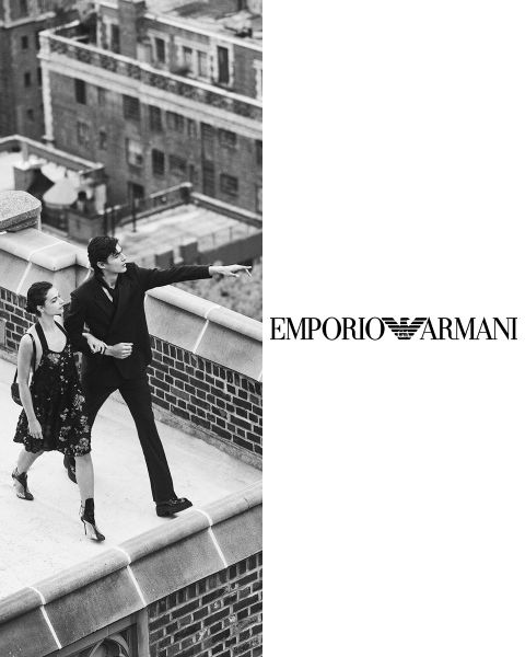 Ein Schwarzweißbild zeigt einen Mann im Anzug und eine Frau im Kleid, die auf einem Dach spazieren gehen. Der Mann zeigt in die Ferne. Auf der rechten Seite ist das Logo von Emporio Armani vor einem weißen Hintergrund zu sehen. Im Hintergrund sind Gebäude zu sehen.