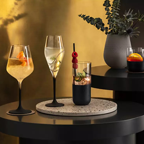 Ein stilvolles Arrangement aus drei eleganten Cocktailgläsern auf einem dunklen Tisch mit einer Keramikvase, in der belaubte Zweige hängen. Jedes Glas enthält ein klares Getränk und Garnituren. Die Szene ist warm beleuchtet und hat einen goldenen Hintergrund.