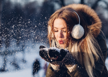 Eine Person mit langen Haaren, die einen Wintermantel mit pelzgefütterter Kapuze und Ohrenschützern trägt, bläst in einer verschneiten Außenumgebung Schnee aus ihren Händen. Der Hintergrund ist verschwommen und zeigt Silhouetten von Menschen und Bäumen.