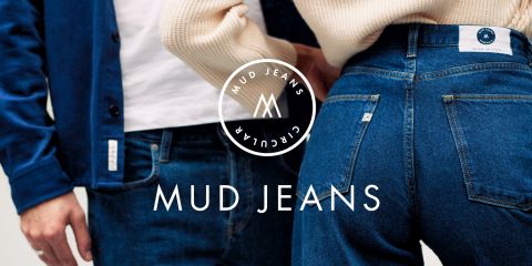 Zwei Personen stehen dicht beieinander und tragen blaue Mud Jeans. Die Person links trägt eine Jacke und die Person rechts zeigt die Rückansicht der Jeans. Das Mud Jeans-Logo und der Text sind deutlich zu sehen.