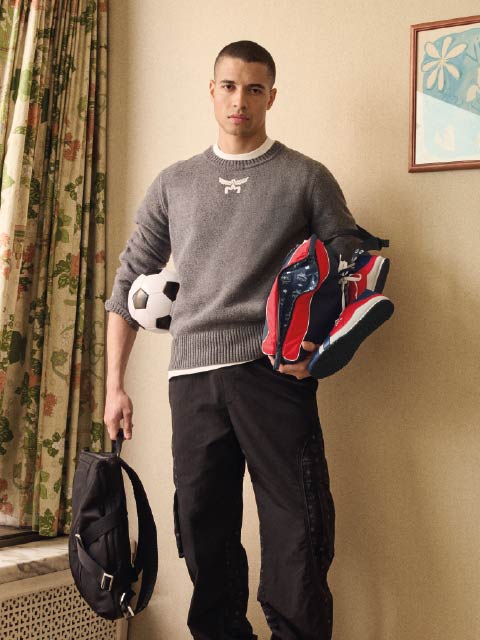 Ein junger Mann in einem grauen Pullover hält einen schwarzen Rucksack und Turnschuhe in der Hand, einen Fußball unter dem Arm, und steht drinnen in der Nähe von geblümten Vorhängen.
