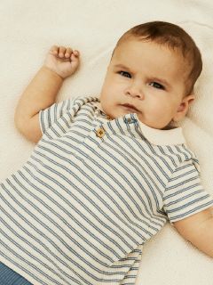 Ein Baby liegt auf einer cremefarbenen Decke und trägt ein gestreiftes Poloshirt. Der Ausdruck des Babys ist nachdenklich, ein Arm ist leicht erhoben und der Kragen ist zugeknöpft. Das Licht ist sanft und warm.