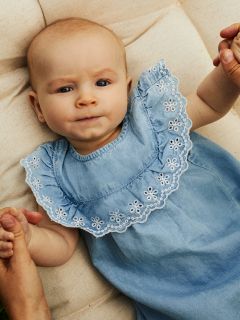 Ein Baby liegt auf einer weichen Oberfläche. Es trägt ein hellblaues Kleid mit aufwendiger weißer Stickerei am Kragen. Das Baby hält die Hände eines Erwachsenen und blickt mit neugierigem Gesichtsausdruck nach oben.