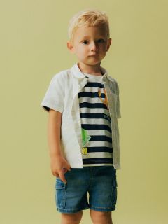 Ein kleines Kind mit blondem Haar steht vor einem hellgrünen Hintergrund. Das Kind trägt ein weißes kurzärmeliges Hemd über einem gestreiften T-Shirt mit grafischem Muster und Jeansshorts. Das Kind hat einen neutralen Gesichtsausdruck.