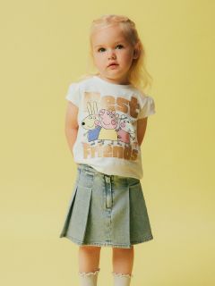 Kleines Kind mit blonden Haaren, trägt ein weißes T-Shirt mit Comic-Tieren und dem Text „Best Friends“ und einen blauen Jeansrock. Der Hintergrund ist hellgelb.