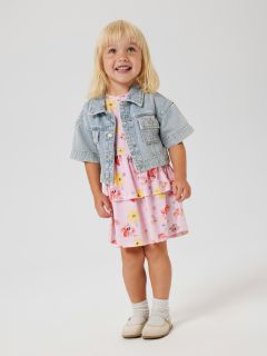 Ein kleines Kind mit blondem Haar steht lächelnd da und trägt eine Jeansjacke über einem rosa Kleid mit Blumenmuster. Es trägt außerdem weiße Socken und helle Schuhe. Der Hintergrund ist schlicht weiß.